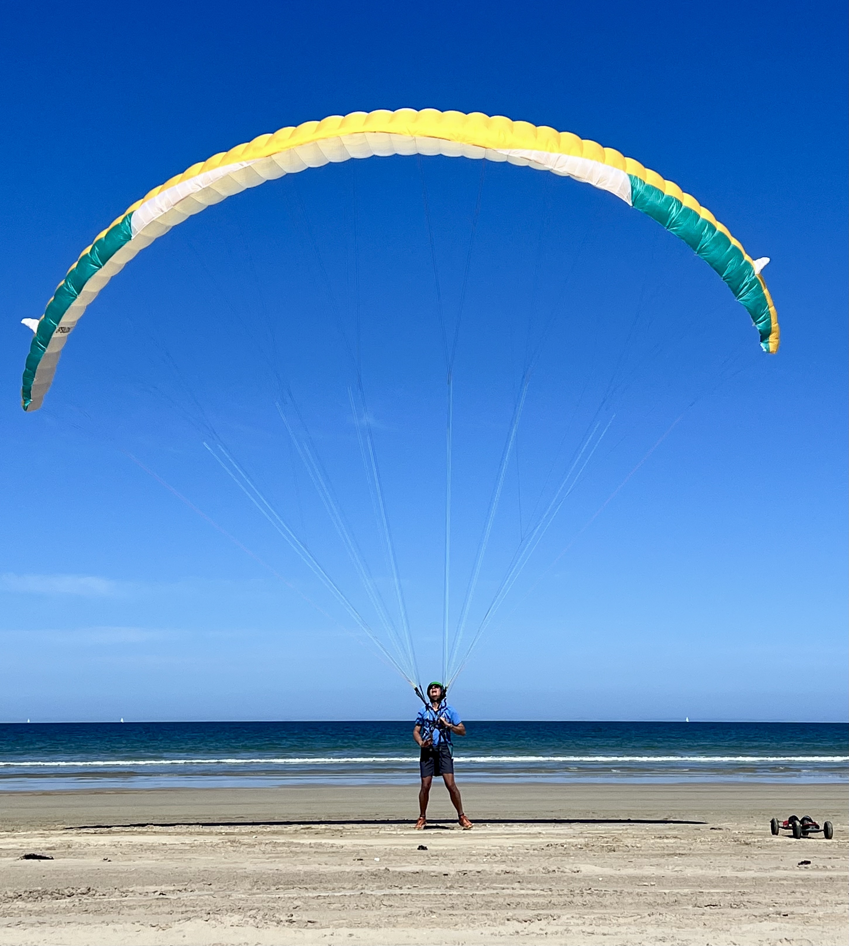Paraglider kiting on beach with yellow and green wing overhead against blue sky