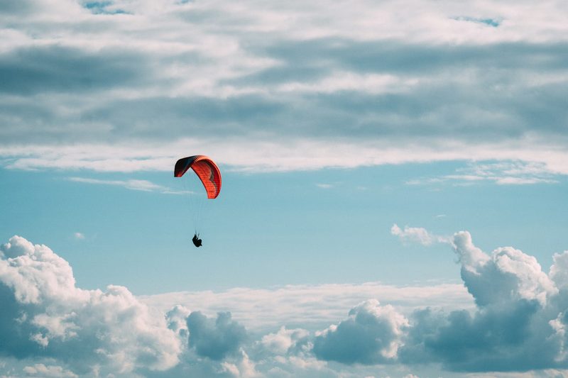 Paraglider with red wing soaring among massive cumulus clouds, showing the vast scale of atmospheric thermals